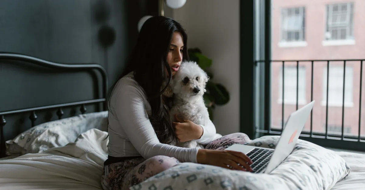 Woman sitting on bed holding a dog, working on a laptop indoors.