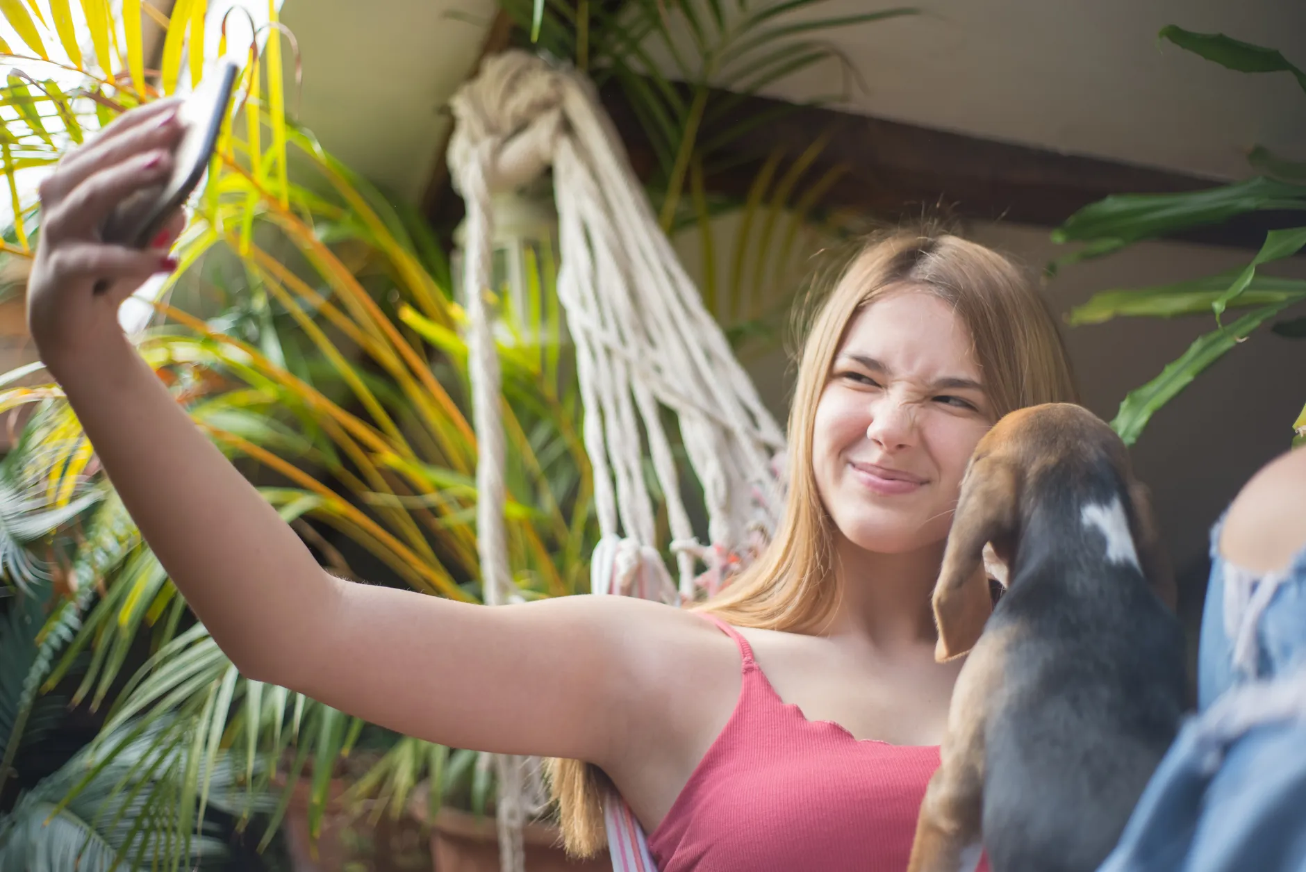 A young woman in a hammock takes a selfie with her playful beagle puppy surrounded by lush greenery.