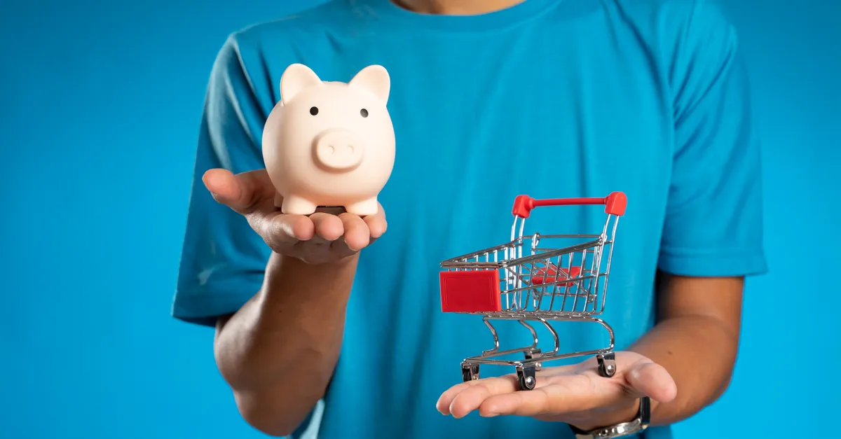 Man in blue shirt holding piggy bank and mini shopping cart, symbolizing savings versus spending.