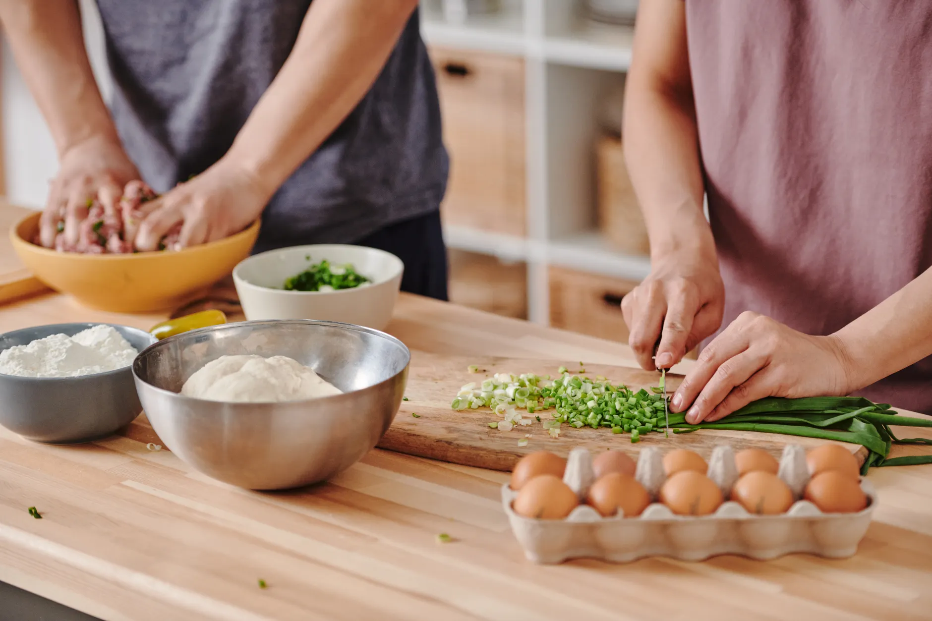 Two people preparing food with fresh ingredients on a wooden table in a cozy kitchen environment.