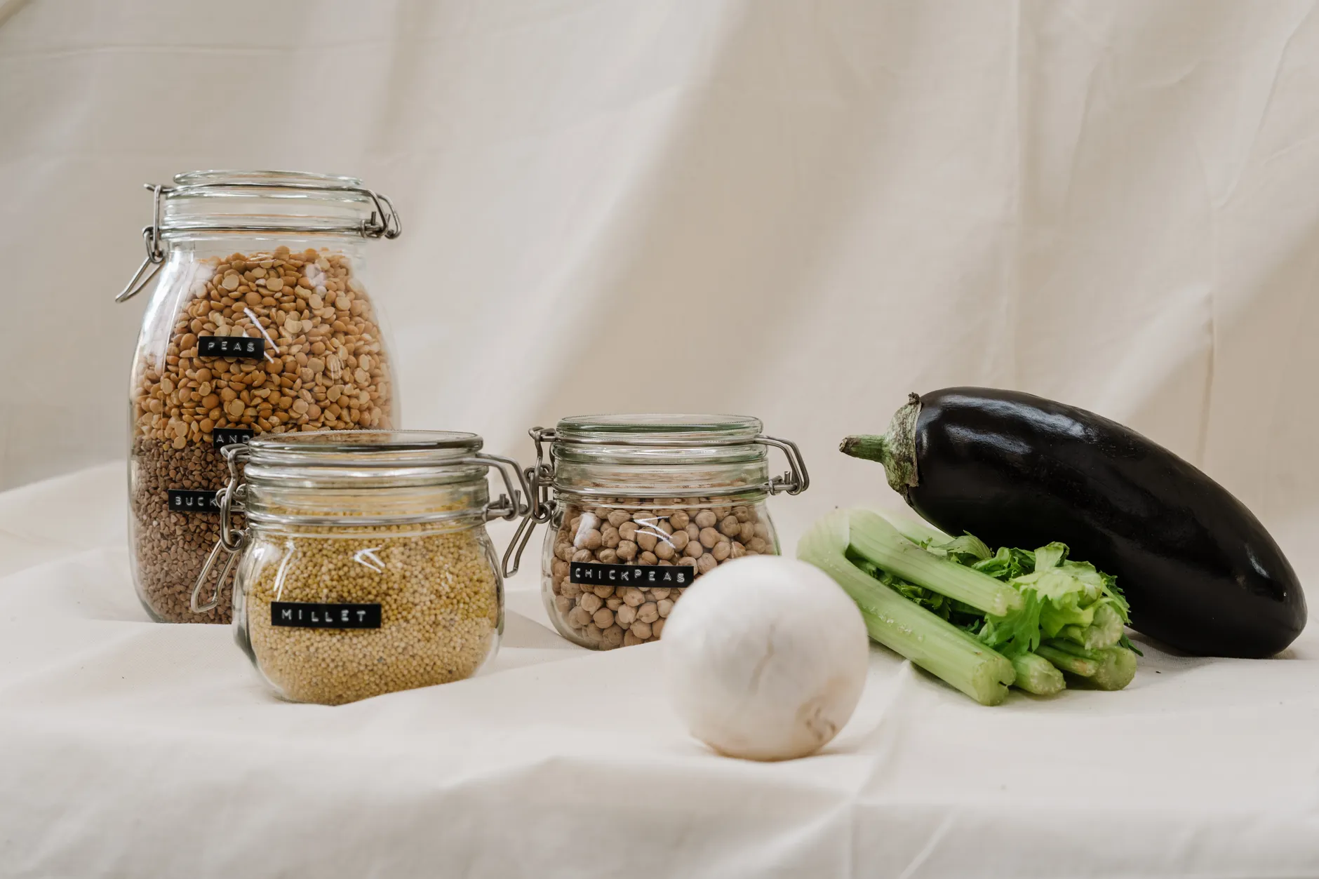 Jars of millet, chickpeas, and vegetables like eggplant and celery on a neutral background.